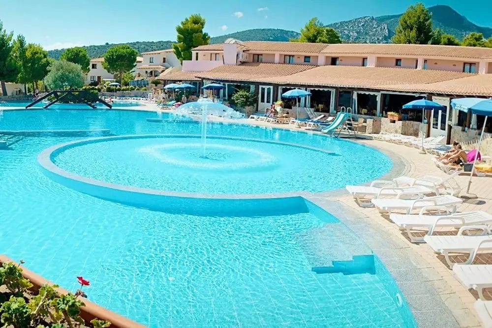 Un villaggio sulla spiaggia con piscina attrezzata con lettini e una bellissima fontana.