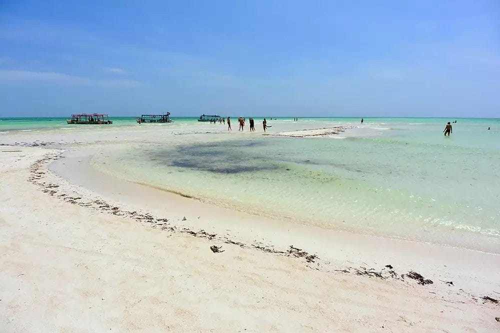 Una spiaggia di sabbia bianca con gente che si gode il sole e barche che punteggiano l'acqua cristallina.