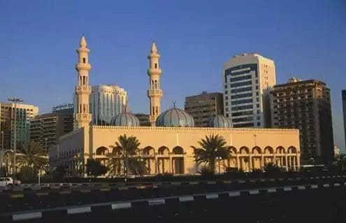 Una grande moschea con minareti gemelli e cupole multiple si erge in primo piano, con moderni edifici urbani, tra cui il ROYAL M HOTEL, che si ergono sullo sfondo sotto un cielo limpido in