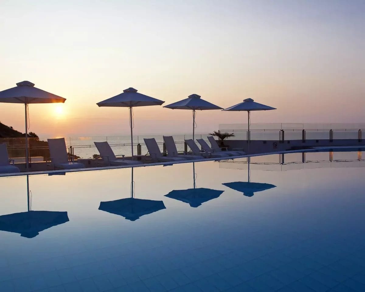 Una piscina con sedie a sdraio e ombrelloni presso l'Apostolata Island Resort & Spa, Cefalonia al tramonto.