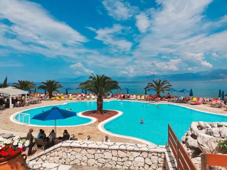 Una vivace scena a bordo piscina in un resort Mareblue Beach con gli ospiti che si godono il tempo soleggiato sotto gli ombrelloni blu.