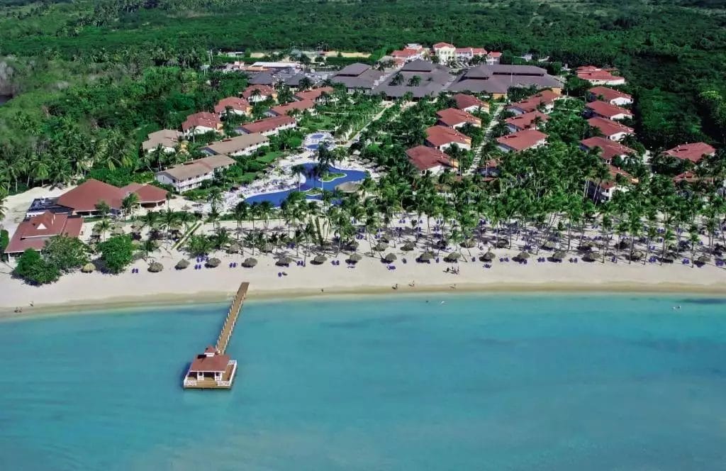 Veduta aerea di un resort sulla spiaggia tropicale con piscine accanto a una tranquilla baia blu con un molo che si estende nell'acqua a Grand La Romana.