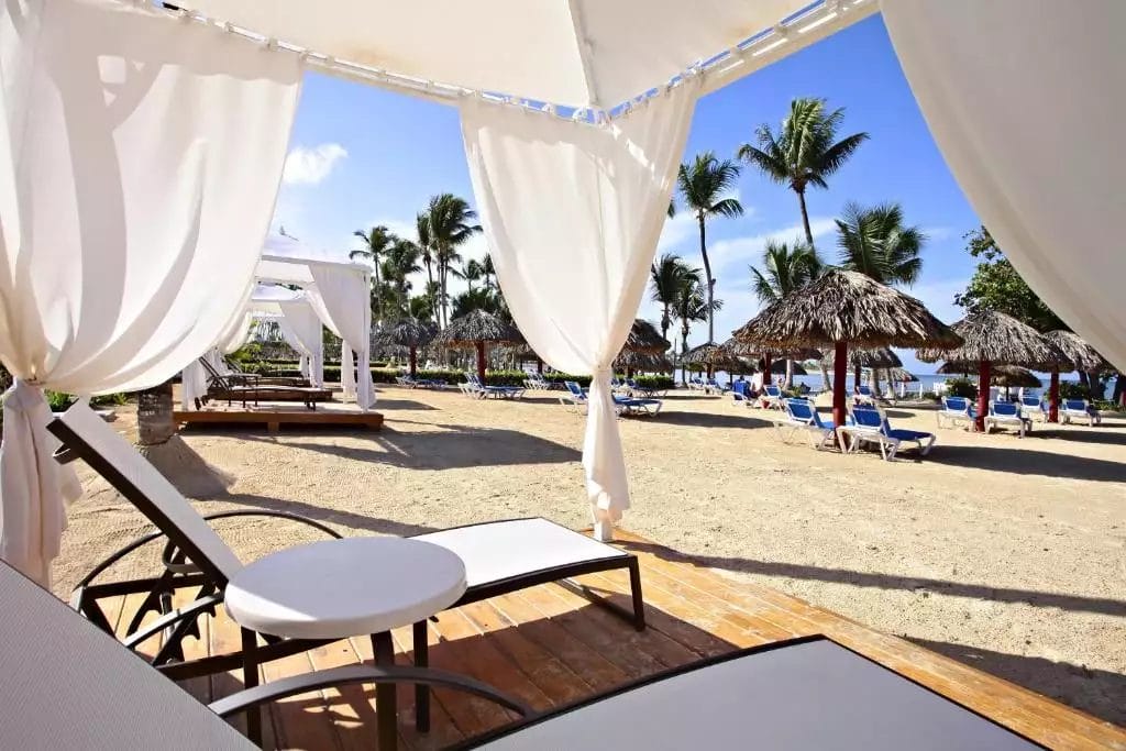 Cabana bianca con tende fluenti al Bahia Principe Grand La Romana, affacciata su una spiaggia sabbiosa con lettini e ombrelloni di paglia sotto un cielo azzurro e limpido.