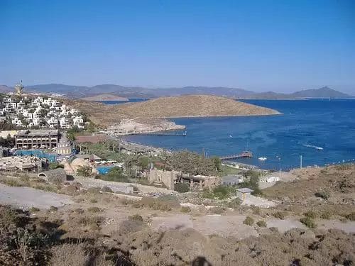 Una vista panoramica sulla costa che mostra gli edifici bianchi del Mare Bodrum Beach Resort appollaiati su una collina che si affaccia su una tranquilla baia blu con un'aspra isola in lontananza sotto un cielo limpido.