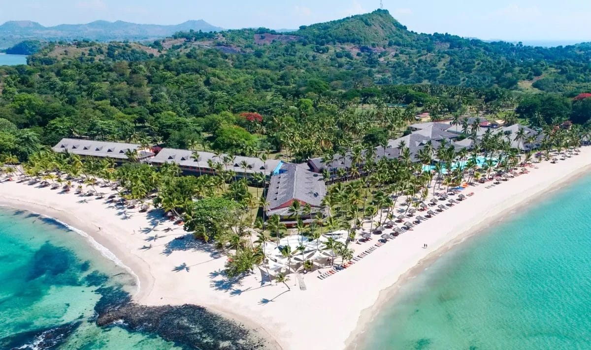 Veduta aerea del Bravo Andilana Beach Resort con ombrelloni e palme lungo una costa curva.