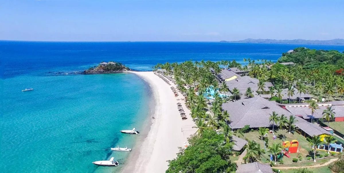 Un resort BRAVO ANDILANA BEACH con palme, edifici dal tetto di paglia e barche vicino alla costa, affacciato su un mare cristallino.