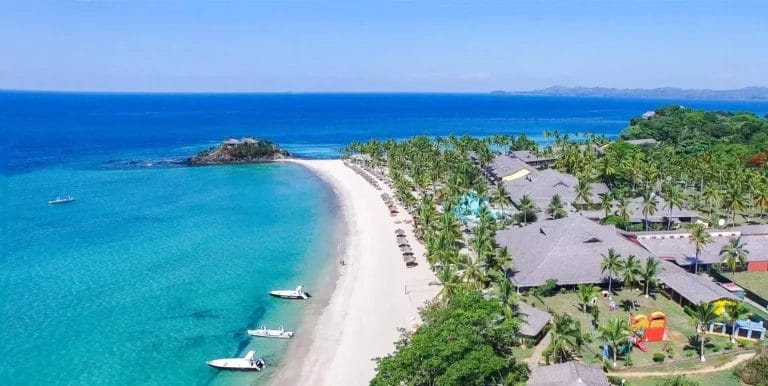 Veduta aerea del resort BRAVO ANDILANA BEACH con palme, tetti di paglia e barche vicino alla riva.