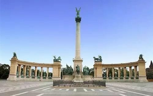 Un maestoso monumento con un pilastro centrale sormontato da una statua, fiancheggiato da colonnati con sculture, sotto un cielo limpido durante un tour di Praga.