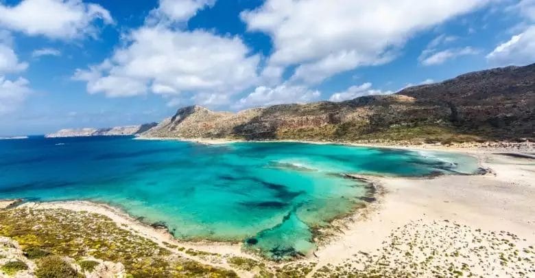 Una vista panoramica di una spiaggia tranquilla con acque turchesi limpide incastonata tra aspre colline sotto un cielo parzialmente nuvoloso, caratteristica del VERAclub Cretan Village.