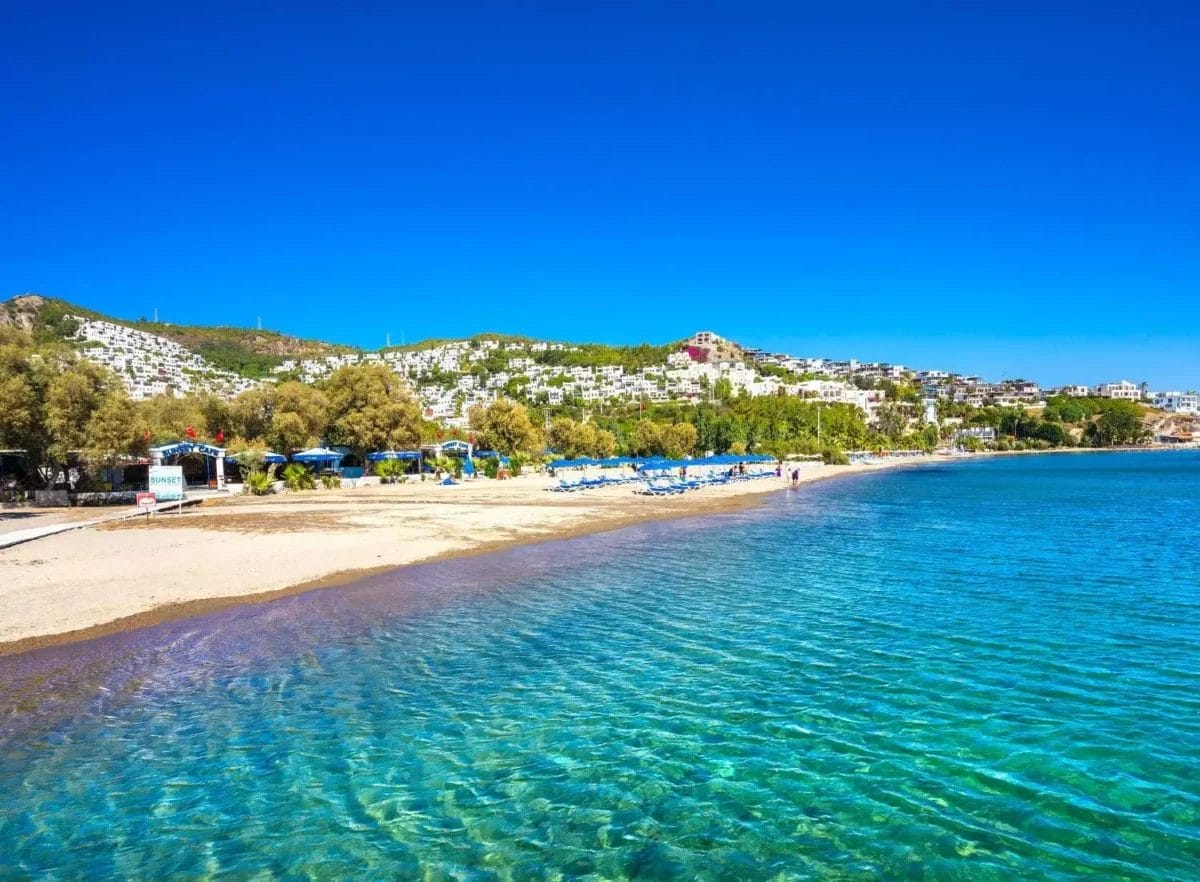 Una spiaggia tranquilla lungo un mare azzurro e cristallino con lettini e ombrelloni allineati sulla sabbia, sullo sfondo di una lussureggiante collina punteggiata di edifici bianchi sotto un cielo azzurro brillante in