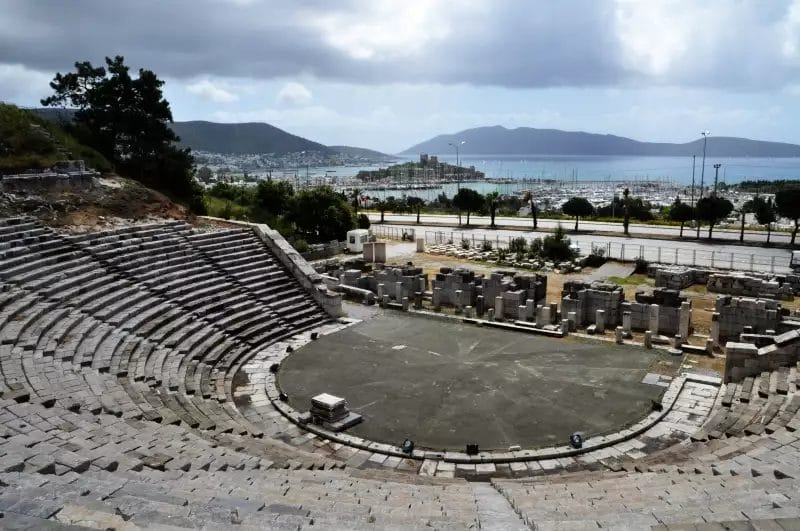 Un antico anfiteatro con posti a sedere in pietra si affaccia su un moderno paesaggio urbano e sul porto della Turchia sotto un cielo nuvoloso.