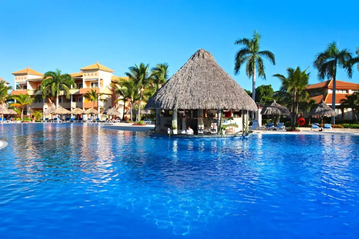 La piscina di un resort tropicale con bar dal tetto in paglia e lettini prendisole sotto il cielo azzurro e limpido del GRAND BAHIA PRINCIPE TURQUESA.