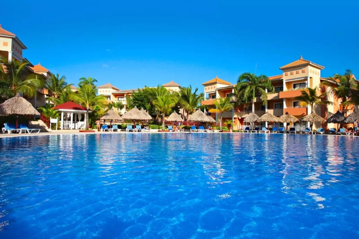 Un resort per vacanze tropicali con una grande piscina in primo piano ed edifici colorati con ombrelloni di paglia sullo sfondo sotto un cielo azzurro.