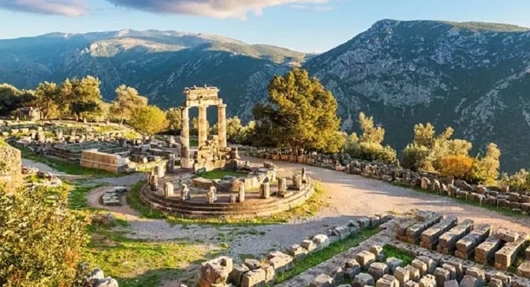 Antiche rovine si crogiolano nella luce dorata del tramonto, con i maestosi resti di una struttura circolare della Grecia classica che si stagliano in mezzo a blocchi di pietra sparsi, il tutto incastonato in uno sfondo lussureggiante