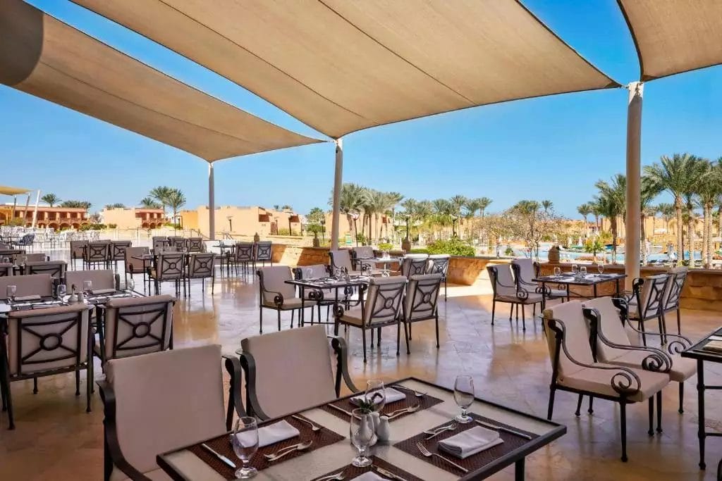 Un'elegante sala da pranzo all'aperto al JAZ GRAND MARSA con mobili eleganti sotto una grande tettoia, che offre una vista su una località soleggiata con palme.