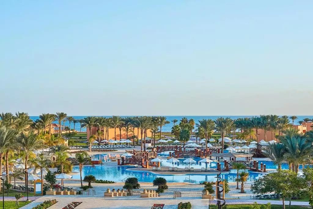 Una tranquilla oasi del resort JAZ GRAND MARSA con palme che circondano una tranquilla piscina, guidando lo sguardo verso il calmo mare blu e il cielo in lontananza.