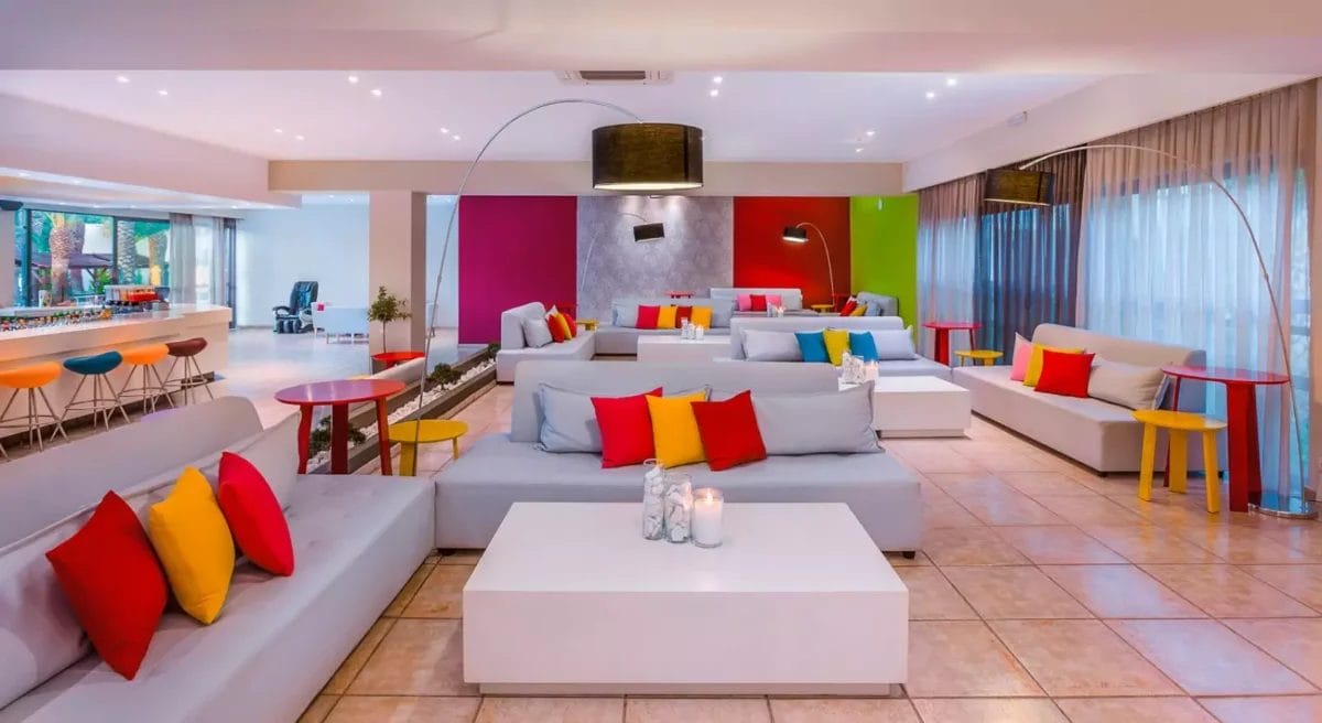 Un'area lounge colorata e moderna nell'hotel KIPRIOTIS HIPPOCRATES con divani bianchi, cuscini rossi e gialli e un bar sullo sfondo.