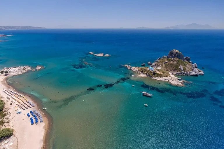 Veduta aerea del paesaggio dell'ALPICLUB AKTI Beach Club con limpide acque turchesi, una spiaggia fiancheggiata da ombrelloni e una piccola isola rocciosa.