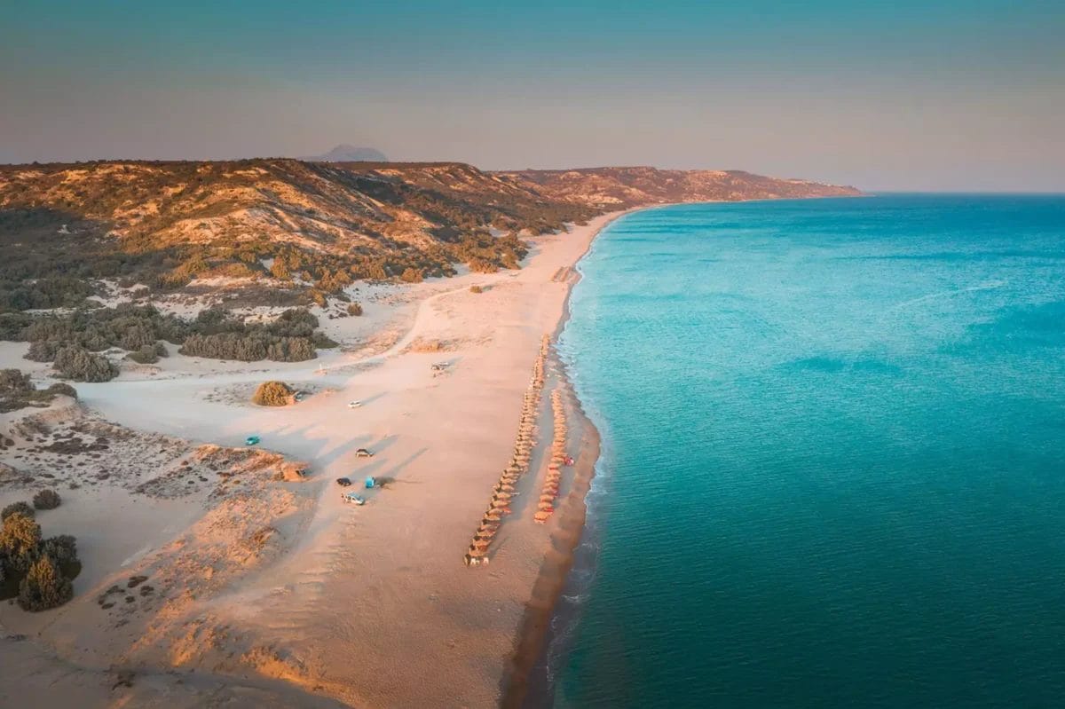 Veduta aerea di una spiaggia tranquilla con file di lettini, adiacente al mare cristallino dell'hotel KIPRIOTIS HIPPOCRATES, durante il tramonto.