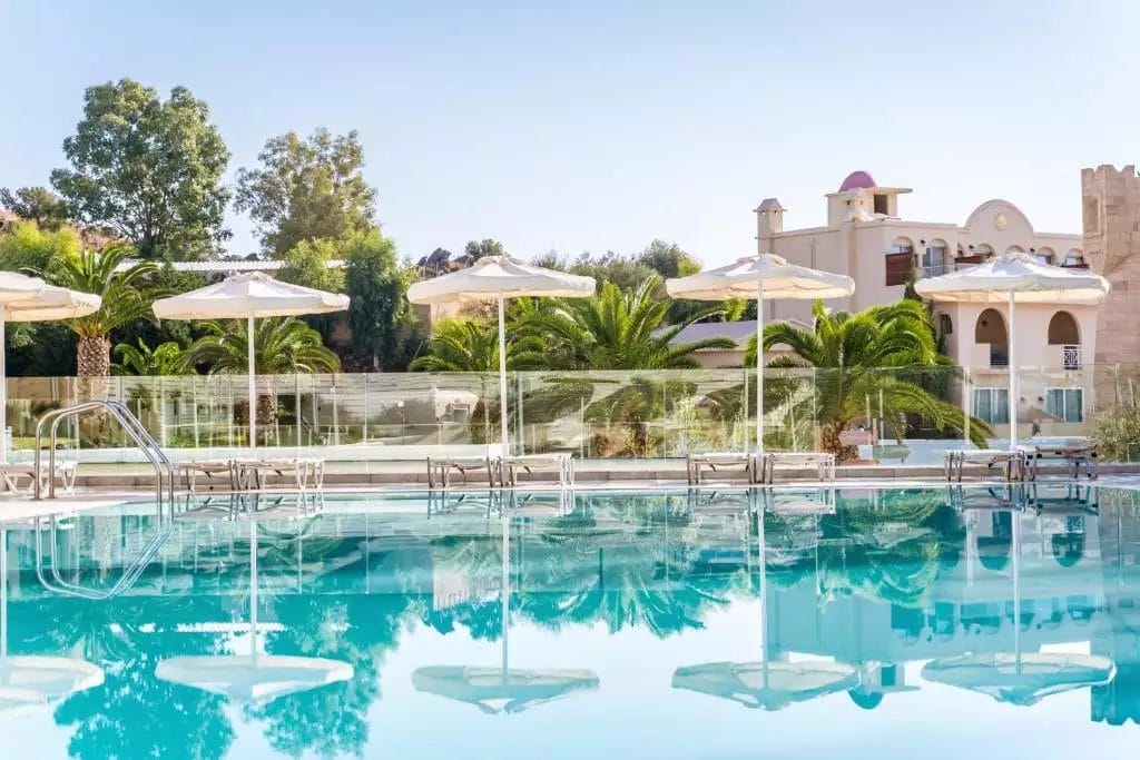 L'ambiente tranquillo a bordo piscina con ombrelloni e architettura in stile mediterraneo sullo sfondo, evidenzia il fascino del Bravo Lindos Royal Resort.