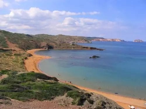 Un pittoresco paesaggio costiero con una spiaggia sabbiosa, calme acque di aguamarina e una collina verdeggiante sotto un cielo parzialmente nuvoloso.