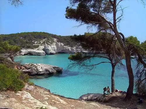 Acque di Aguamarina di una baia tranquilla incorniciata da scogliere rocciose e ombreggiata da verdi pini, con persone sulla riva.