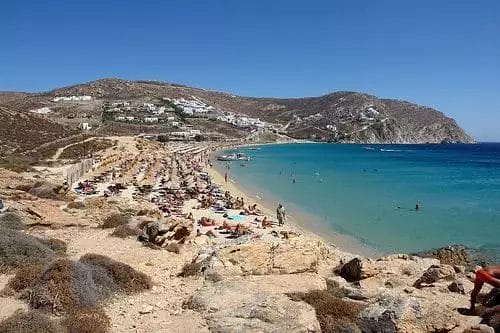 Un affollato Paradise Beach Resort con bagnanti e acque cristalline, delimitato da terreno accidentato ed edifici bianchi sul fianco della collina.