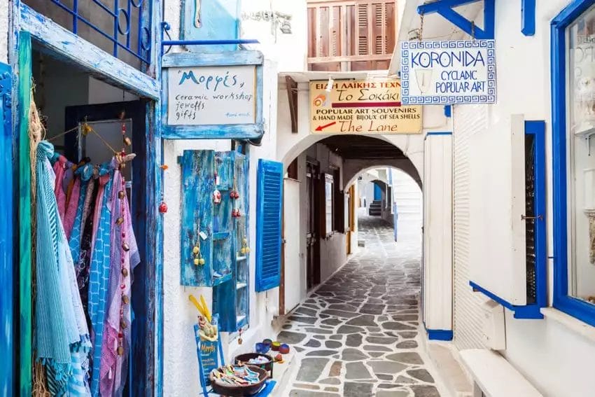 Vicolo stretto a VERACLUB NAXOS, una destinazione turistica con pareti bianche, porte blu e negozi locali che espongono prodotti colorati.