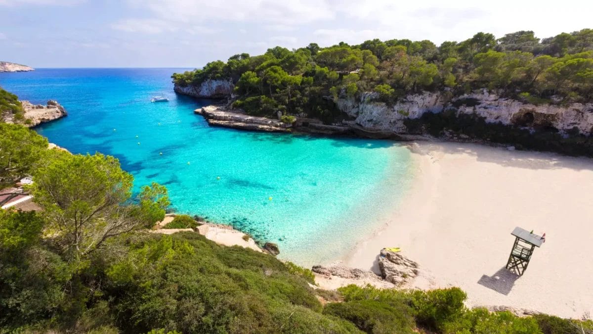 Veduta aerea di una tranquilla insenatura di MAIORCA con acque turchesi cristalline, circondata da scogliere e vegetazione mediterranea, caratterizzata da una spiaggia di sabbia bianca e una torre di salvataggio.