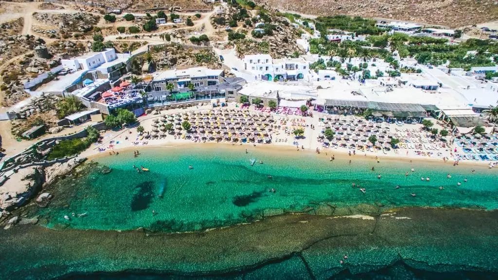 Veduta aerea di un affollato Paradise Beach Resort con acque turchesi, adiacente ad edifici bianchi e lettini allineati lungo la riva.