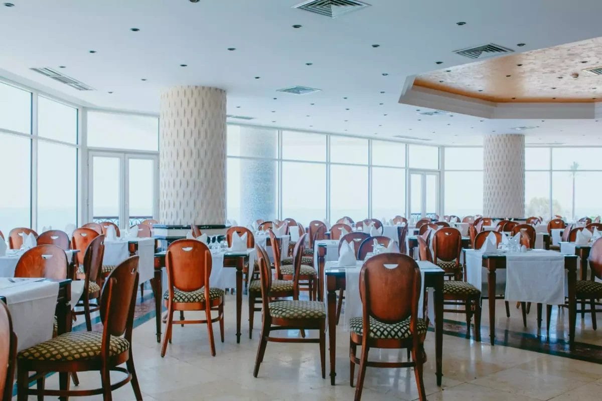 Un'elegante sala da pranzo all'interno del Queen Sharm Resort con grandi finestre che consentono ampia luce naturale, con tavoli rotondi vestiti con tovaglie bianche e circondati da eleganti sedie in legno con rivestimento rosso, il tutto contrapposto a un