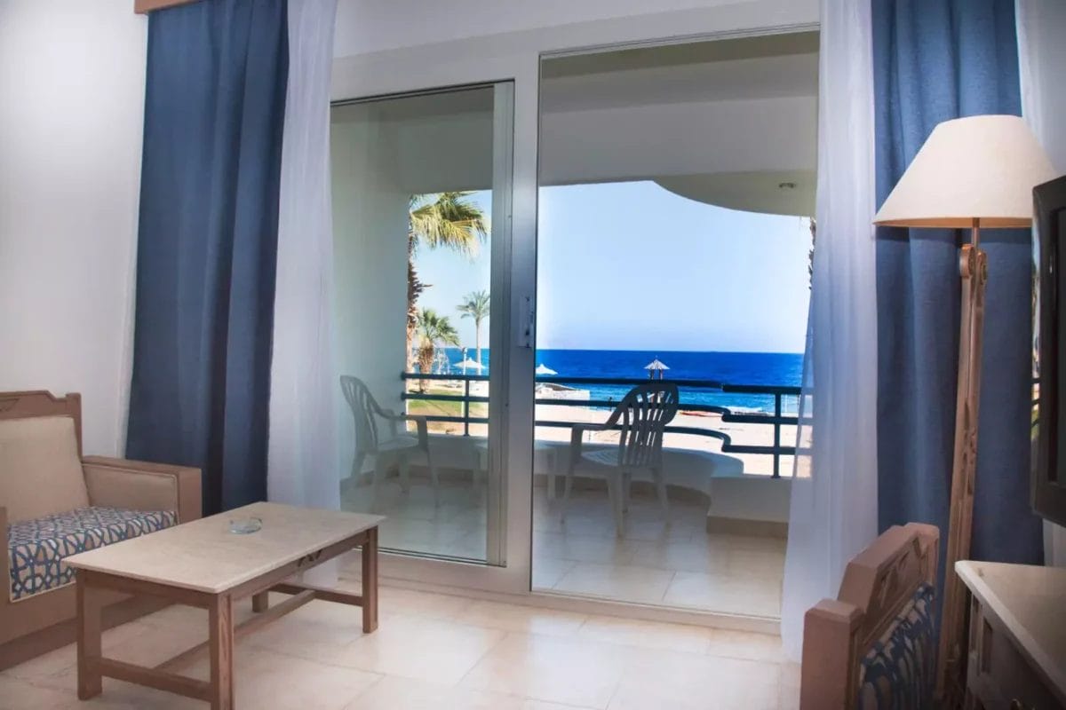 Una serena vista della costa dal balcone della camera d'albergo del Queen Sharm Resort, che invita al relax con viste del mare blu profondo e del cielo limpido, incorniciati da morbide tende blu scuro.