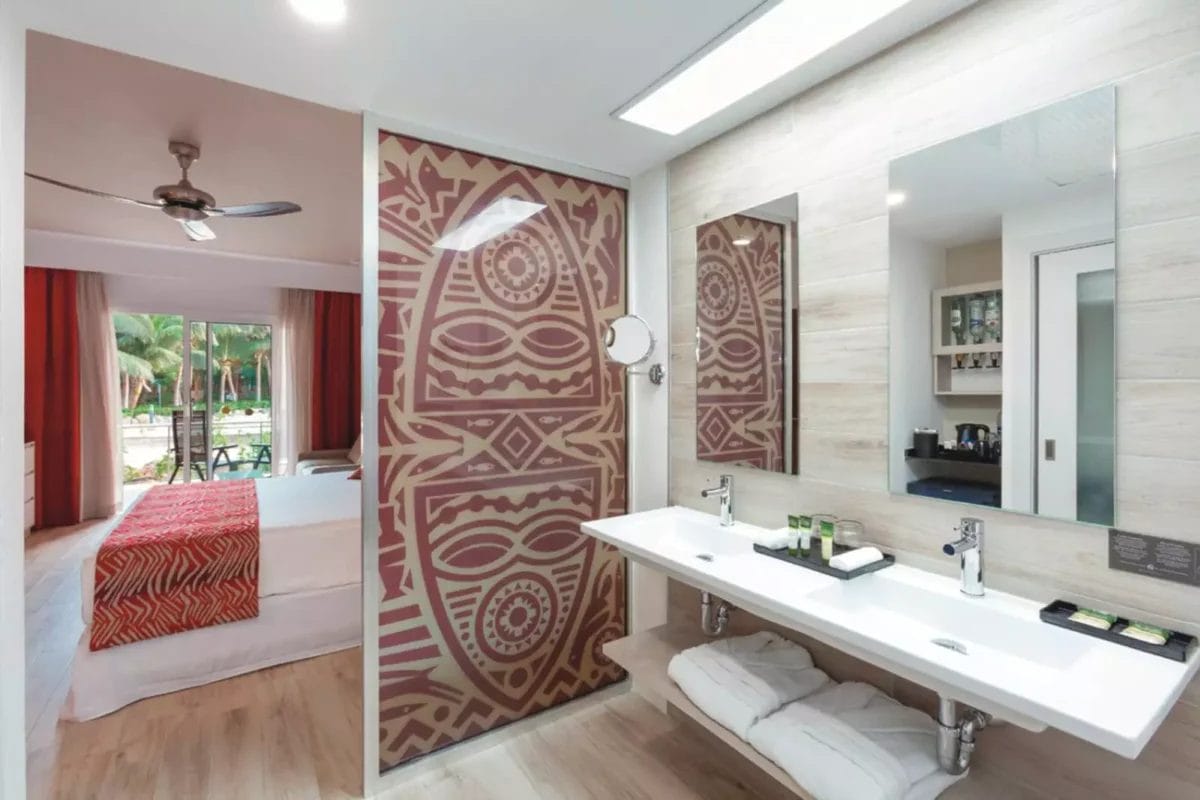 Bagno moderno con doppie porte scorrevoli decorate con motivi geometrici, che conduce ad una camera da letto con vista sulle palme nel resort RIU Cabo Verde.