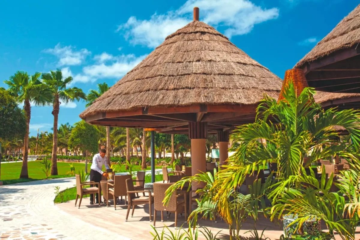 Ambiente tropicale da resort RIU Cabo Verde con padiglioni dal tetto di paglia e un cameriere che serve un tavolo.