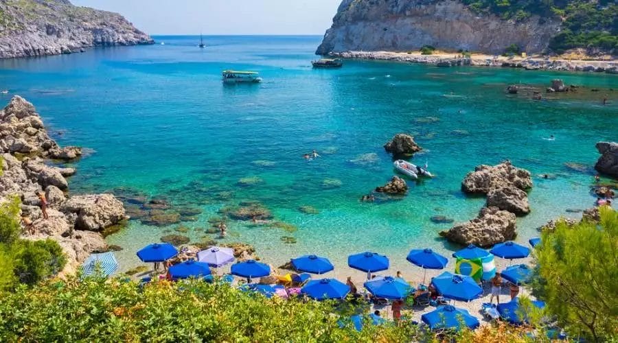 Un pittoresco litorale al Lindos Royal Resort con persone che nuotano, ombrelloni blu che punteggiano la spiaggia e barche che galleggiano nell'acqua cristallina.