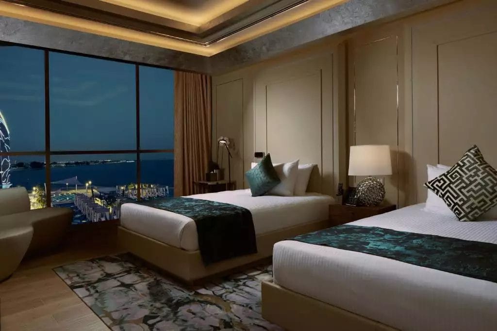 Una lussuosa camera presso il Royal M Hotel di Abu Dhabi con un arredamento moderno, dotata di due letti con biancheria da letto elegante, arredi eleganti e una grande finestra che offre una vista mozzafiato del paesaggio urbano notturno e