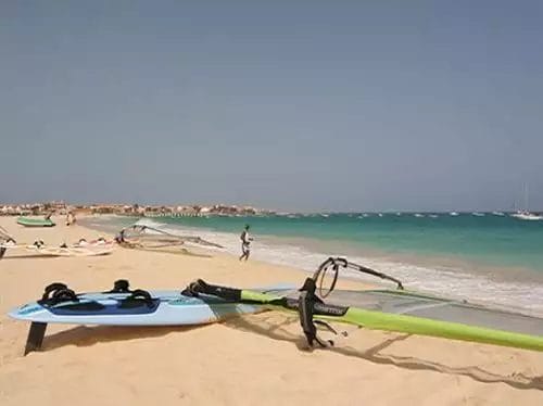 Una spiaggia sabbiosa del RIU Cabo Verde con tavole da surf e attrezzatura da windsurf, affacciata su un mare calmo con bagnanti in lontananza.