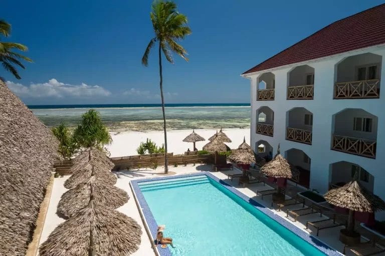 Una persona che nuota nella piscina del resort Sun Bay vicino alla spiaggia, con palme e ombrelloni sotto un cielo limpido.