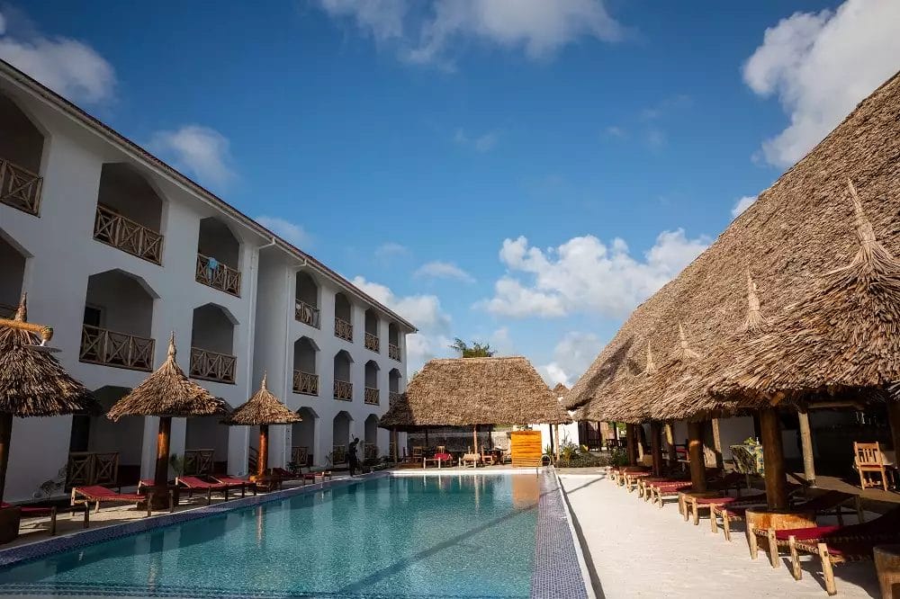 Un resort tropicale con piscina fiancheggiata da edifici bianchi e cabine dal tetto di paglia sotto un cielo azzurro e limpido, situato a Sun Bay.