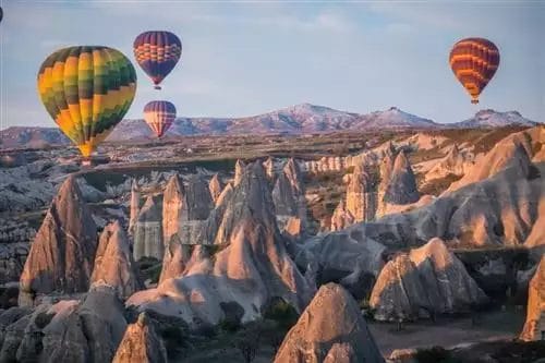 Mongolfiere colorate sorvolano la spiaggia di Mare Bodrum, caratterizzata da formazioni rocciose uniche sotto un cielo tenue e pastello.