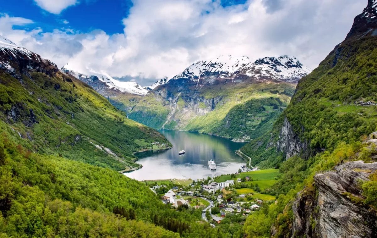 Una vista panoramica di un fiordo in Norvegia con un villaggio annidato tra rigogliosi pendii verdi e montagne innevate sullo sfondo.