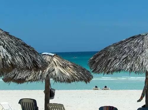 Gli ombrelloni di paglia forniscono ombra su una soleggiata spiaggia di Varadero con acque cristalline e alcune persone che si rilassano sui lettini in lontananza.