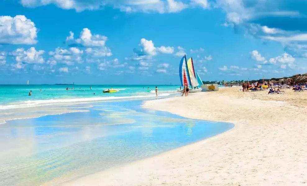 Una scena soleggiata sulla spiaggia di Varadero con acque limpide e poco profonde sulla costa, una barca a vela colorata sulla destra e persone impegnate in attività in spiaggia sotto un cielo parzialmente nuvoloso.