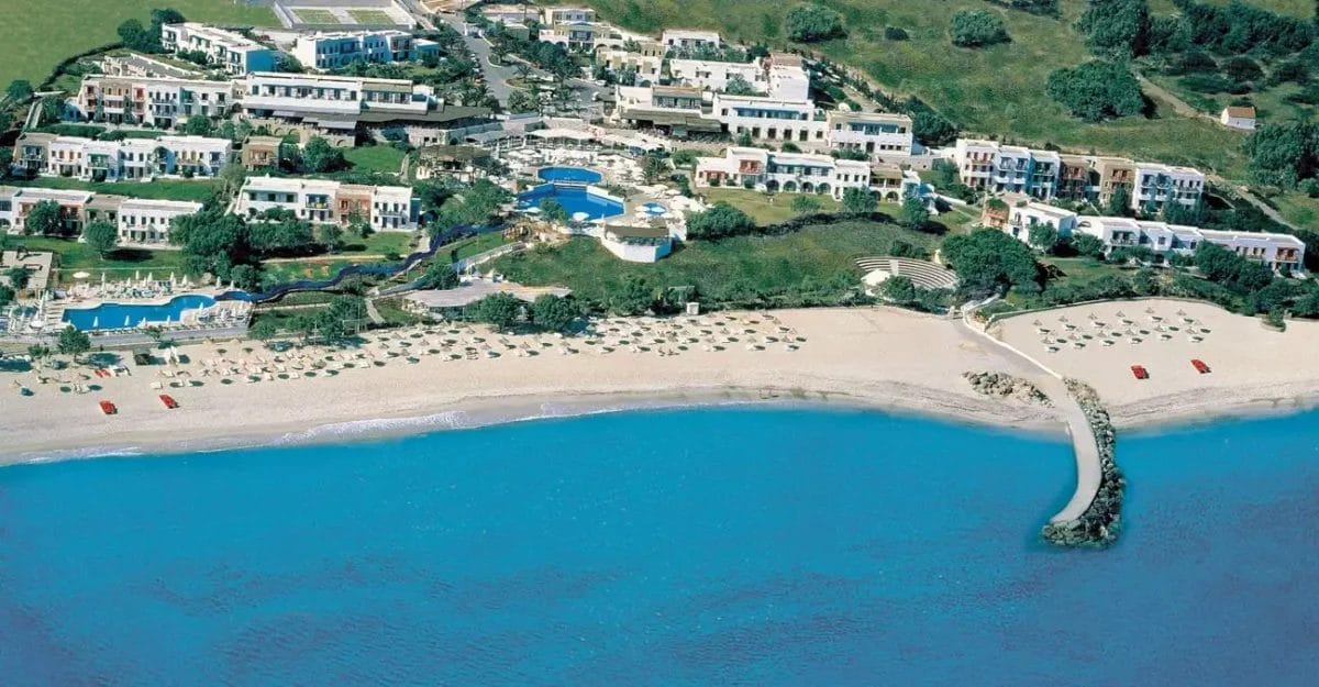 Resort costiero con edifici bianchi e piscine adiacenti ad una spiaggia sabbiosa, immerso in un pittoresco villaggio cretese.