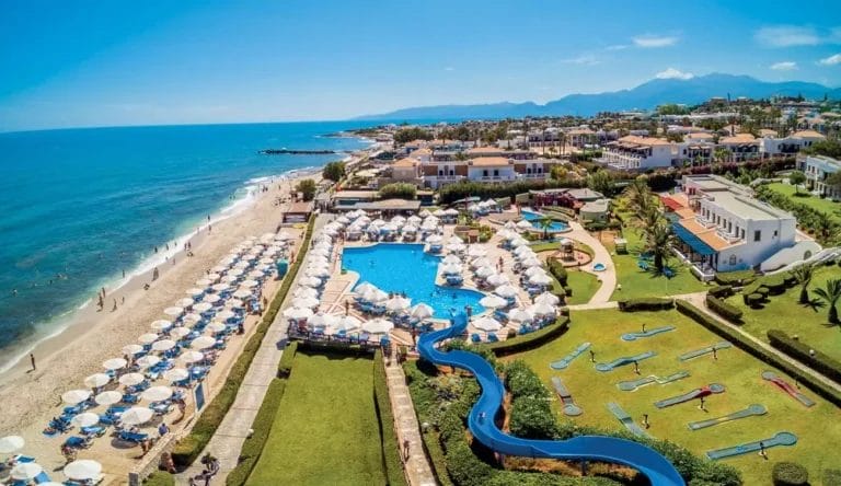 Veduta aerea di un resort costiero del villaggio cretese con piscina, ombrelloni ed edifici circostanti.