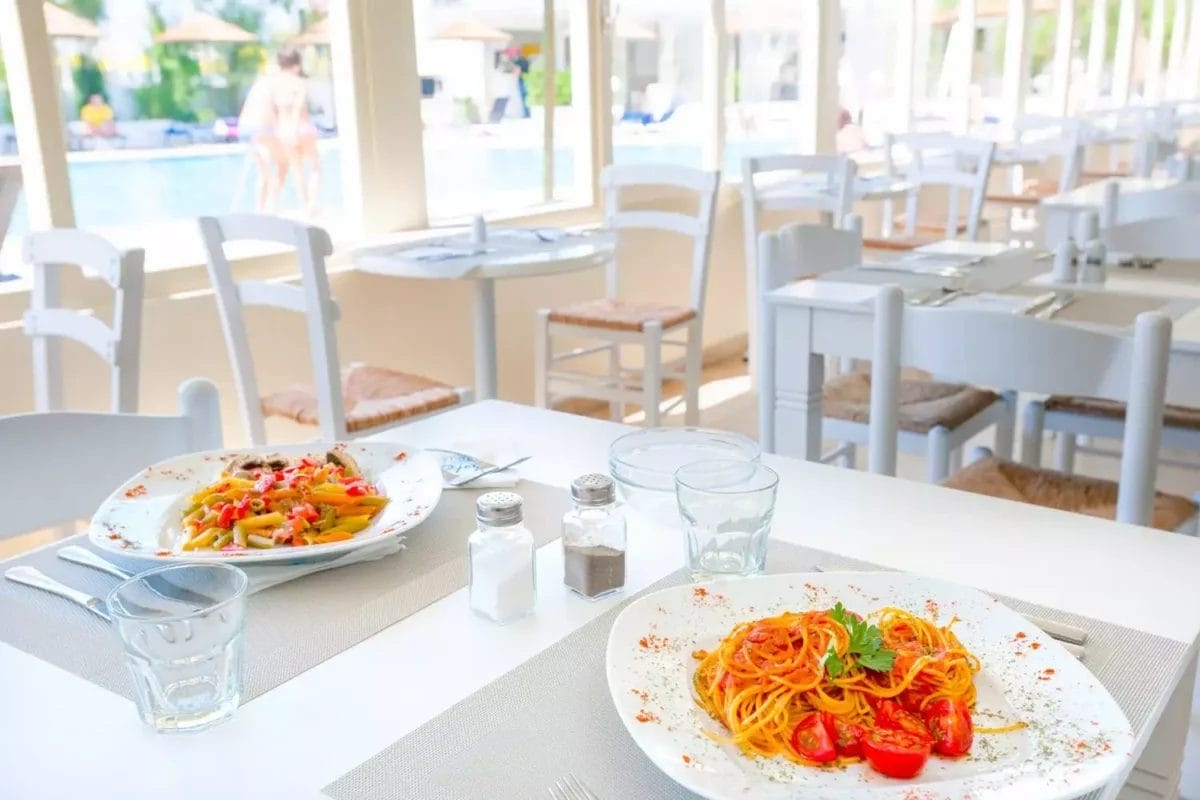 Interni luminosi e moderni del ristorante con piatti di pasta serviti su tavoli bianchi, con vista sull'area piscina del VERACLUB NAXOS.