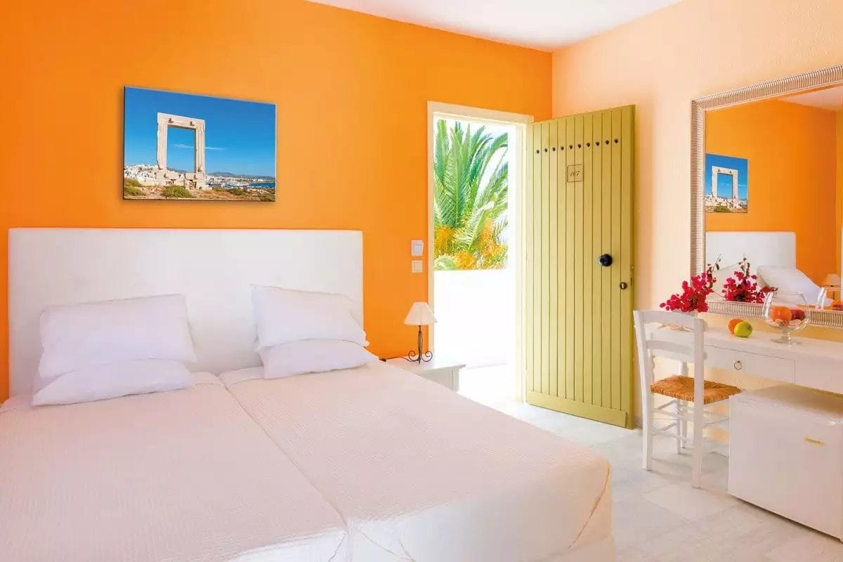 Una camera da letto luminosa e invitante con biancheria da letto bianca, una parete arancione e un vivace arredamento in stile mediterraneo ispirato al VERACLUB Naxos.