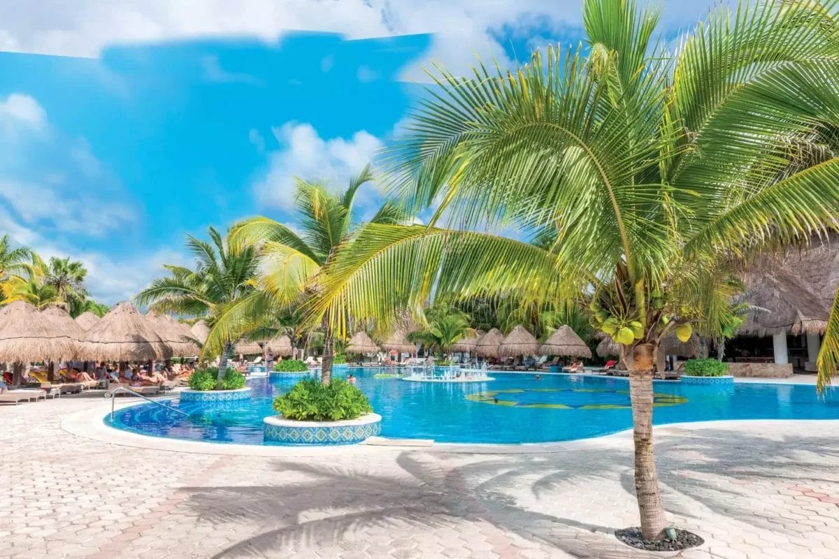 Il bordo piscina del Royal Tulum Resort con palme e ombrelloni in paglia sotto un cielo azzurro.