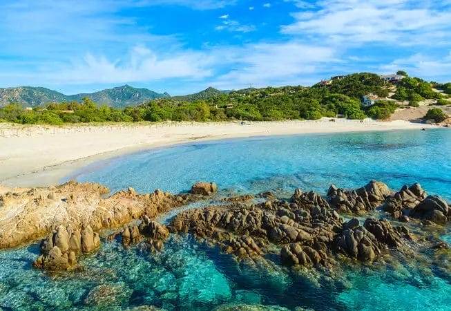 Tranquilla spiaggia sabbiosa del VOI Tanka Resort con acque turchesi cristalline, rocce frastagliate in primo piano e vegetazione lussureggiante sullo sfondo sotto un cielo azzurro brillante.