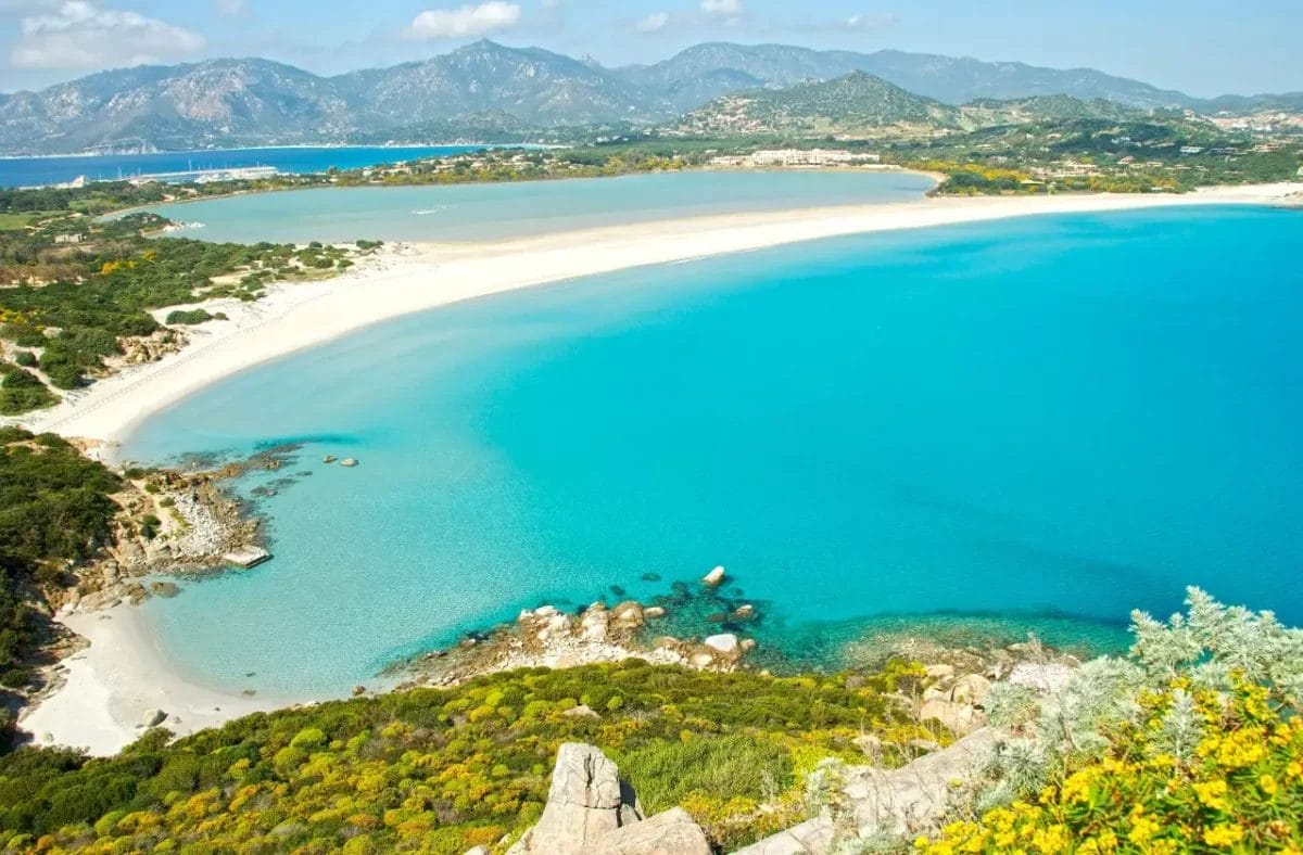 Una vista panoramica di un sereno paesaggio costiero della Sardegna con acque turchesi cristalline, un'ampia spiaggia sabbiosa vicino al VOI Tanka Resort e una vegetazione lussureggiante sotto un cielo soleggiato.